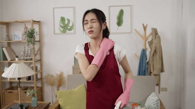 asian cleaning woman suffering a sudden crick in her neck is groaning and exercising the muscles while using a hoover for chores in the living room at home