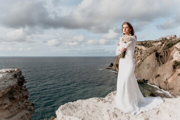 Fototapeta na wymiar Romantic bride, blond girl in white wedding dress with open shoulders posing with sea and rocks in background. Stylish young woman standing on cliff's edge and looking into a ocean mountains at sunset