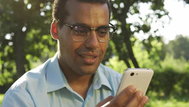 Happy positive biracial man smiling typing on smartphone, betting online in special app and winning, free spins and moves, real money or cryptocurrency, digital way to earn money