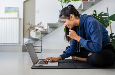 Young Hindu woman sitting on a mat with a laptop preparing a yoga class. Exercise at home concept