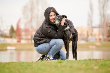 Ground level of smiling young woman in outerwear with dark hair looking at camera while hugging loyal black dog while kneeling on lawn on gray day in park.