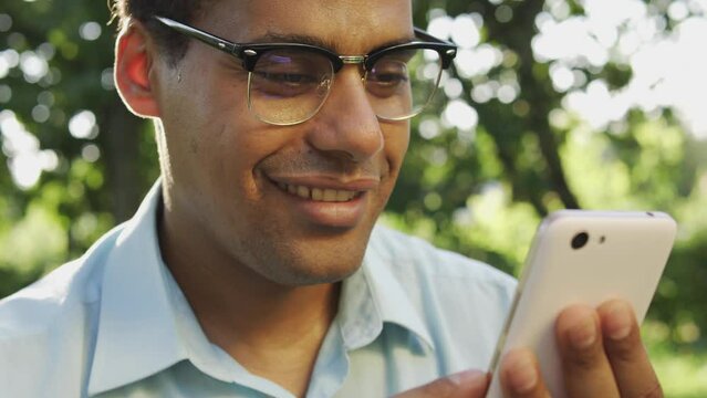 Smiling biracial man in eyeglasses scrolling web-pages on phone, shopping online