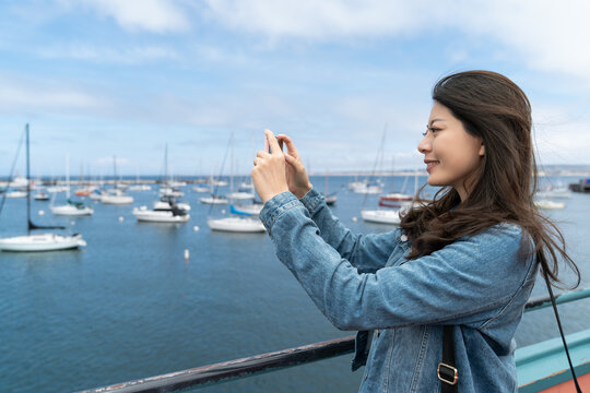 side portrait of cheerful asian chinese girl tourist taking photos of sailing fishing boat on blue ocean with her smartphone while traveling at Old Fisherman's Wharf in Monterey California usa