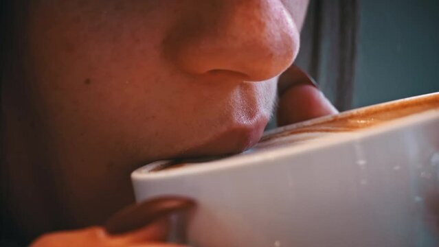 Young woman drinks coffee with foam close-up. Hot cappuccino with foam is brought to the lips. A girl takes a sip of hot latte in a cozy coffee shop.