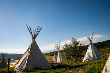 View of teepees on grassy field with blue sky in background. Tents for camping on landscape during...