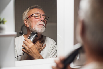 Senior man is trimming his beard with electric trimmer in bathroom.