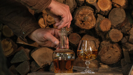 Cognac pouring in the glass from bottle. Hand puts a carafe with golden cognac and pouring into the glass, wooden table near logs. Strong drink - brandy or whiskey with ice. Alcohol amber drink