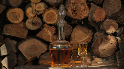Cognac pouring in the glass from bottle. Hand puts a carafe with golden cognac and pouring into the glass, wooden table near logs. Strong drink - brandy or whiskey with ice. Alcohol amber drink