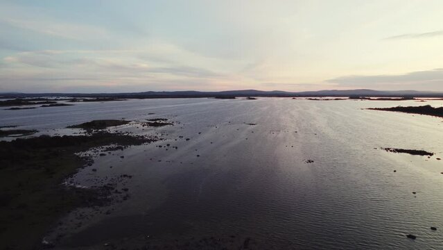 Beautiful aerial flying over gigantic irish lough corrib with many small island silhouettes in the water. Flying forward, over part of coast during blue hour golden evening sun reflecting in waves