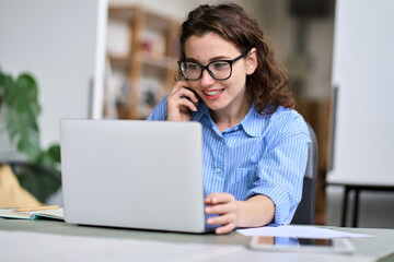 Young woman sales manager talking on cell phone using laptop working online on computer. Female...