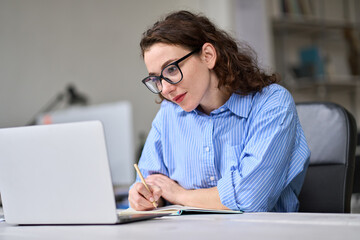 Young business woman employee using laptop writing notes, having remote virtual work conference meeting call in office or watching webcast online webinar training web course, doing research.