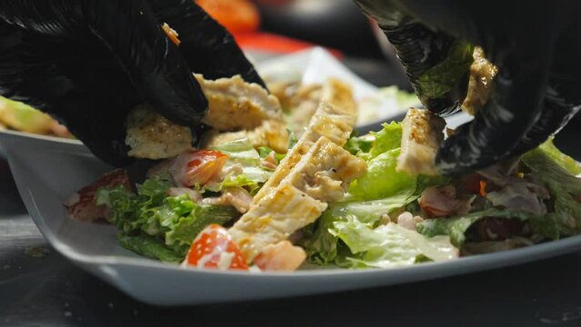 Male arms putting pieces of fried chicken unto fresh ingredients in white plate at cuisine. Hands of chef in gloves arranging caesar salad for being served. Concept of cooking food. Close up Slow mo