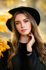 Close up portrait of a Beautiful girl in a dark dress and black hat standing near colorful autumn leaves. Art work of romantic woman .Pretty tenderness model looking at camera.