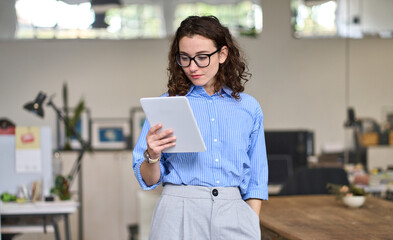 Young smart busy professional business woman executive, female company worker or manager holding digital tablet using pad technology device working standing in modern corporate office.