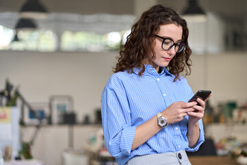 Fototapeta na wymiar Busy young business woman manager holding smartphone working standing in office using mobile cell phone, checking corporate apps, browsing online on cellphone typing on cellular technology device.