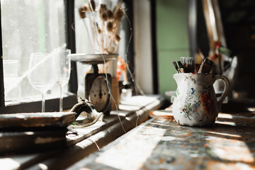handmade jug with paintbrushes near vintage scales and glasses on windowsill in sunlight.