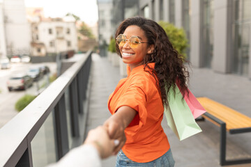 Smiling young black woman with packages with purchases leads friend by hand, rejoices to good...