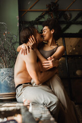 Fototapeta na wymiar sexy interracial artists hugging and kissing near vase with dried plants in workshop.