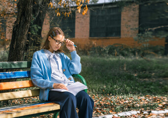 A teenager on a bench is doing homework on a sunny autumn day.