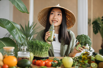 Beautiful asian woman in traditional conical hat holding broccoli and smoothie sitting at table with fruit and vegetables ingredients to make healthy breakfast, indoor at stylish tropical kitchen.