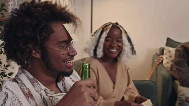 Young Black man with sticker on forehead holding bottle of beer playing funny guessing game at party with friends