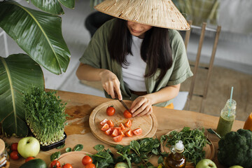 Beautiful asian young woman in traditional conical hat is slicing cherry tomatoes, sitting at table in bright exotic studio with many fresh fruits and vegetables are preparing for cooking.