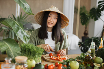 Beautiful asian young woman in traditional conical hat is slicing cherry tomatoes, sitting at table in bright exotic studio with many fresh fruits and vegetables are preparing for cooking.