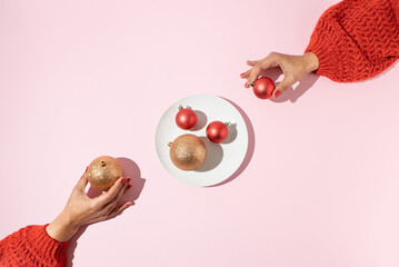 Two female hands taking Christmas baubles of the white plate on pink background. Minimal holiday...