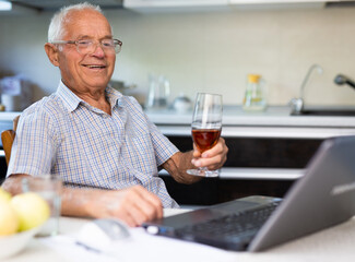 Fototapeta na wymiar Happy elderly man with glass of wine at laptop sitting at table