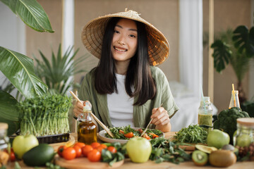 Happy young beautiful asian woman making healthy salad stirring chopped leaves indoor at exotic...