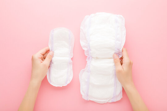 Young adult woman hands holding opened white sanitary towels on pink table background. Big and small size. Pastel color. Hygiene product for urinary incontinence or after childbirth. Top down view.