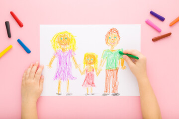 Baby girl hands drawing happy family with colorful wax crayons on light pink table background....