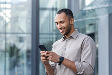 hispanic man outside modern office building using smartphone, businessman in shirt typing message and browsing online pages.