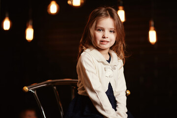 Little adorable smiling girl is sitting on metal chair with a dark background with retro light...
