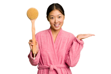 Young asian woman holding a shower brush isolated showing a copy space on a palm and holding another hand on waist.