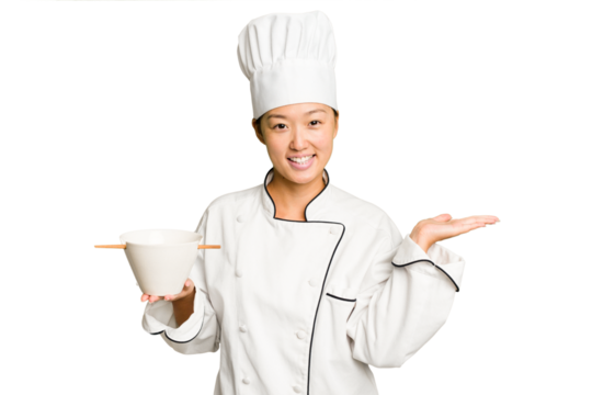 Young chef asian woman holding a bowl of ramen isolated showing a copy space on a palm and holding another hand on waist.