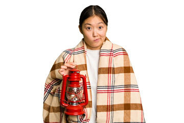 Young asian woman wearing a blanket and holding a lamp isolated shrugs shoulders and open eyes...