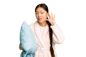 Young asian woman wearing a pajama holding a pillow isolated trying to listening a gossip.