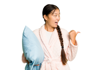 Young asian woman wearing a pajama holding a pillow isolated points with thumb finger away, laughing and carefree.