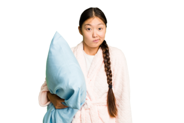 Young asian woman wearing a pajama holding a pillow isolated shrugs shoulders and open eyes confused.