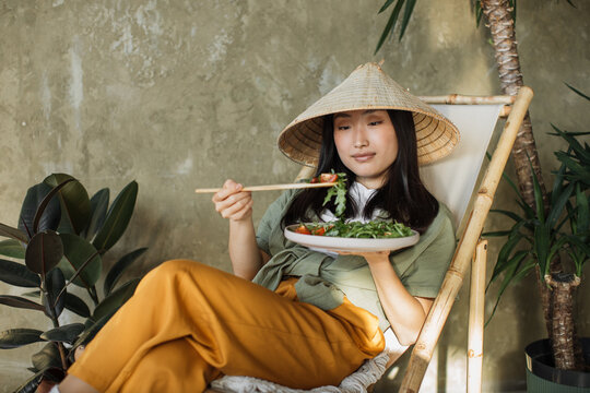 Portrait of young asian woman holding vegetarian salad while sitting on folding chair with organic vegetables, enjoying healthy diet, indoor at grey background. Weight loss concept.