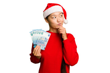 Young asian woman celebrating Christmas holding banknotes isolated looking sideways with doubtful and skeptical expression.