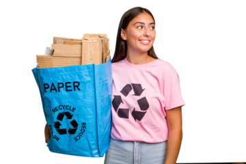 Young caucasian woman holding a recycling bag full of paper to recycle isolated looks aside smiling, cheerful and pleasant.