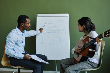 Side view of confident teacher of music explaining musical notes to student with acoustic guitar...