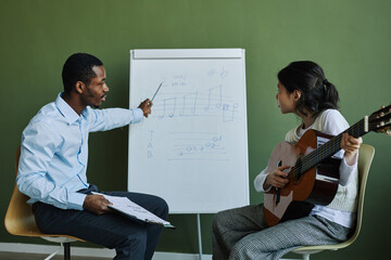 Side view of confident teacher of music explaining musical notes to student with acoustic guitar while pointing at whiteboard