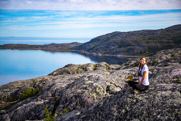 Naklejka na ściany i meble girl sits on rocks admiring the sunrise over a bay in northern norway; hiking in nordic norway, rugged landscape of the norwegian fjords