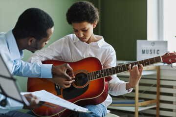 Youthful diligent girl playing acoustic guitar while sitting in front of African American male...