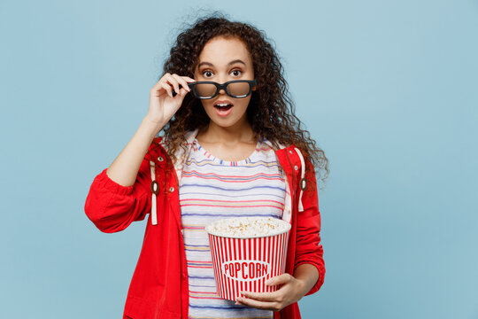 Young shocked surprised woman of African American ethnicity lower 3d glasses watching movie film hold bucket of popcorn in cinema isolated on plain pastel light blue cyan background studio portrait.