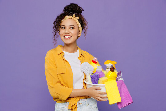 Young fun housekeeper woman wear yellow shirt tidy up hold basin with detergent bottles look aside on workspace area mock up isolated on plain pastel light purple background studio. Housework concept.