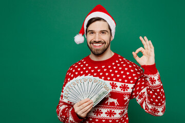 Merry young man wear red knitted sweater Santa hat posing hold in hand fan of cash money in dollar...
